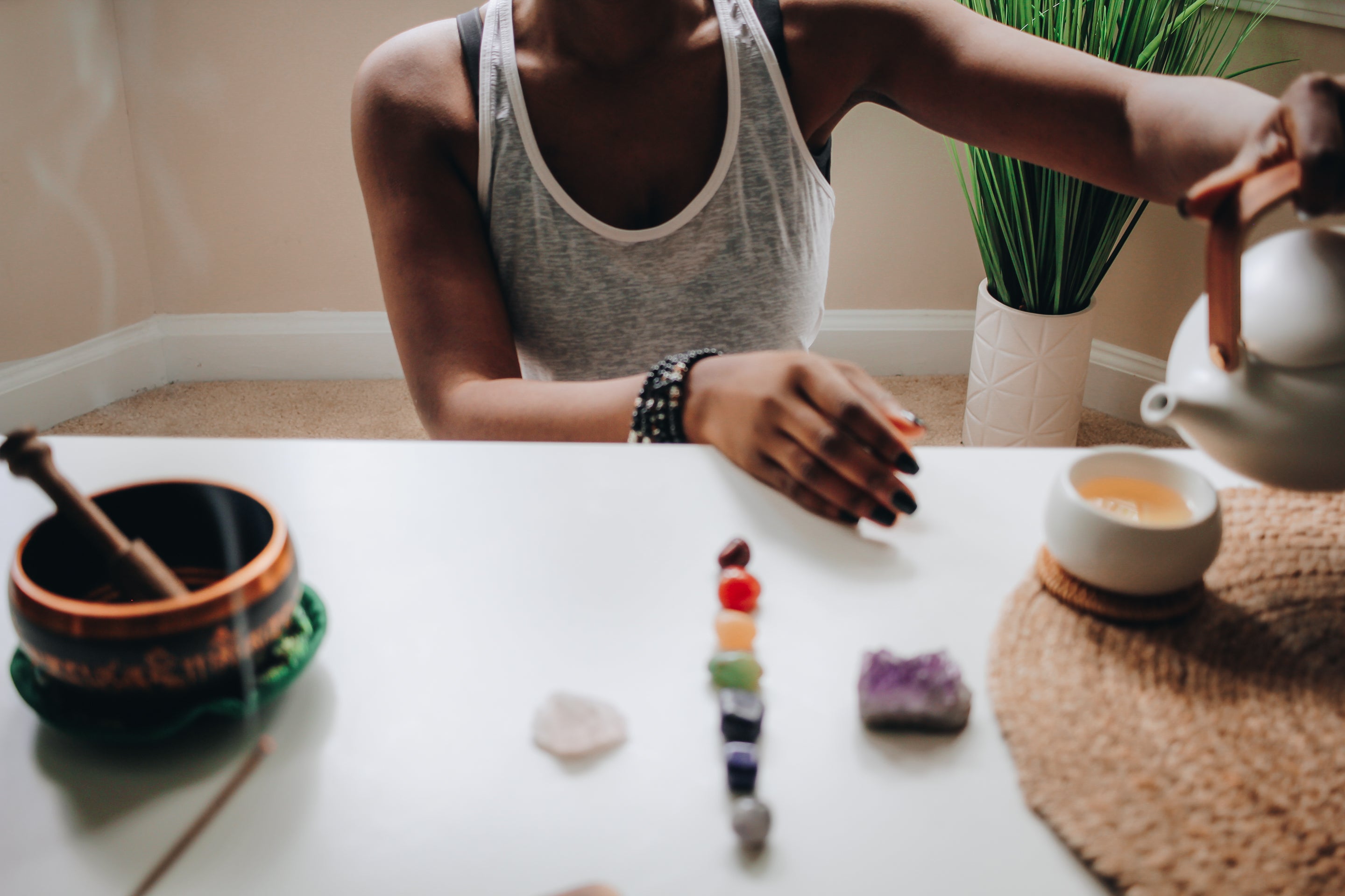 Photograph of woman pouring a pot of tea into a small white cup resting on a bamboo mat. There are stones representing the chakras directly in front of her and to the left is a small singing bowl and a smoldering incense. A plant is also seen in the back.